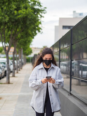 Mujer joven con mascarilla usando y hablando por su teléfono móvil cumpliendo las medidas de...