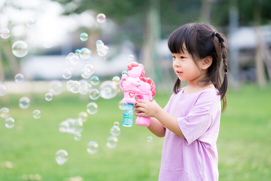 Happy Asian cute girl is playing a pink soap bubble gun. Child have fun playing on the lawn. A 4 year old children play outdoors. Leisure activities.