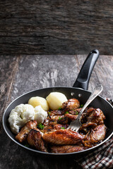 Roasted chicken wings with potato and cauliflower in skillet on wooden table
