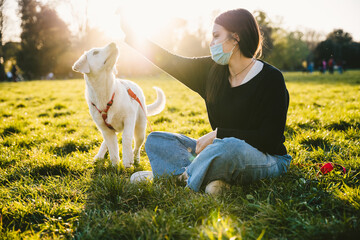 Young woman at the park at sunset plays and teaches behaviors to her dog wearing a face mask against Coronavirus infections, Covid-19 - Millennials having fun with her pet friends