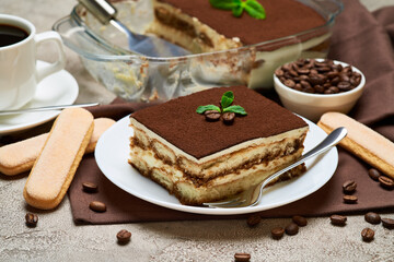 Traditional Italian Tiramisu dessert in glass baking dish and portion on grey concrete background