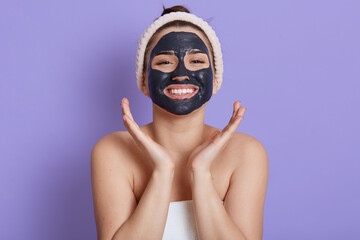 Happy female doing moisturizing procedures for skin of face, looks at camera with smile, keeping palms near cheeks, expressing joy, cosmetology manipulation, isolated over lilac background.