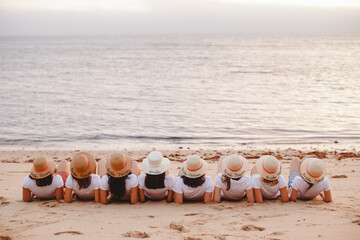 Group of young women sitting on the beach wearing similar panama hats facing the ocean