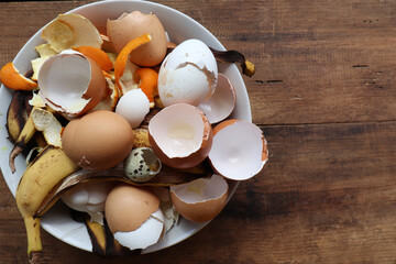 Banana skin, eggshell, and orange peels in white bowl on wooden table background. Using kitchen...