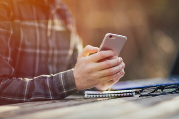Hands of a young man freelancer is typing  message on his smartphone on a wooden table during break time from working on the laptop.
