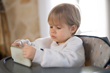 Fototapeta premium Cute little boy using smartphone, looking at screen, curious child holding phone in hands, sitting on a chair at home.