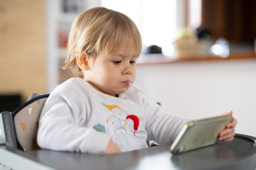 Cute little boy using smartphone, looking at screen, curious child holding phone in hands, sitting on a chair at home.
