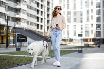 Cheerful woman walking with her big white dog on a leash on the street near residence while have a...