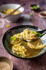 Side view of two bowls of home made vegan wonton soup one spoon with a dumpling held up served on wooden table with chopsticks with chili crisp oil