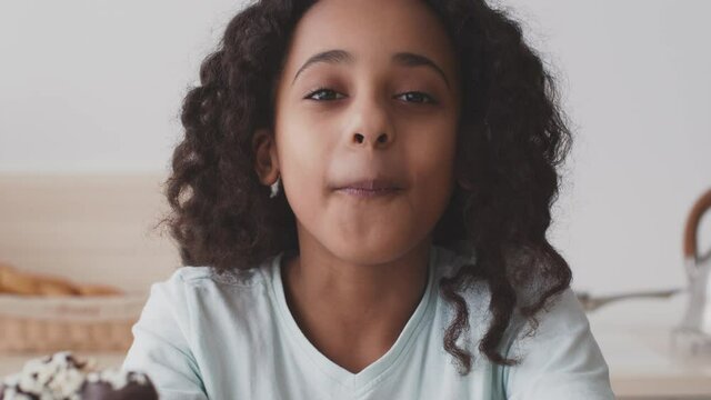 Cute african american girl enjoying sweet chocolate donut, biting dessert and smiling to camera, close up portrait