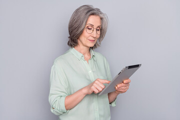 Photo of positive lovely focused concentrated mature woman in glasses using tablet isolated on grey color background