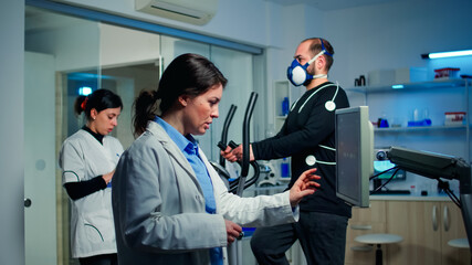 Doctor sport scientist checking EKG scan at monitor and discussing while man with mask running on cross trainer monitoring his endurance. Team of researchers monitoring his endurance and heart rate