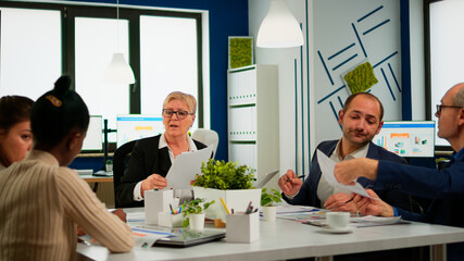 Woman secretary bringing documents and coffee to executive director while multiethnic team planning financial strategy during business conference. Manager briefing teamworkers during brainstorming.