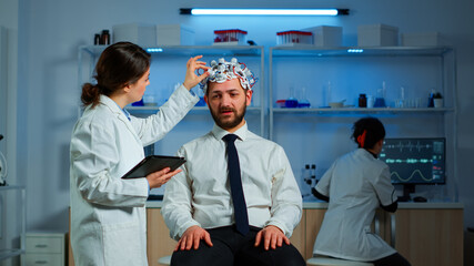Obraz premium Young man wearing brainwave scanning headset discussing with neurological researcher about symptoms of disease. Medical scientist writing informations of brain scan on tablet working in laboratory