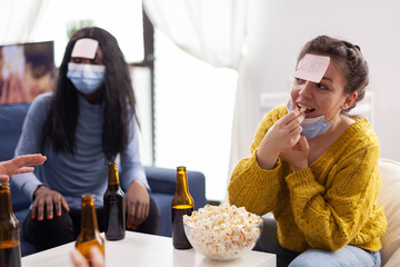 Happy woman with sticky note on forehead playing name game with multiethnic friends keeping social distancing drinking beer in home living room due to social pandemic. People having fun in outbreak