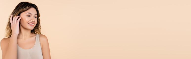 Obraz premium joyful asian woman fixing hair while looking away isolated on beige, banner