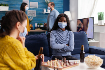 African woman wearing face mask playing chess drinking beer, keeping social distancing preventing spread of covid virus. Diverse people having fun in home living room playing board games