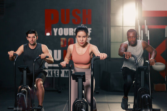 Group Of Determined Multi Ethnic People At Gym Exercising On Air Bike. Concentrated Fitness Asian Woman Training On Exercise Bike With Class Riding Cycling Machine In Hard Effort..