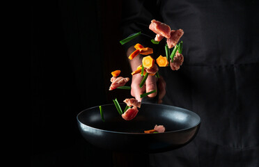 The hand of a professional chef tosses pieces of vegetables and meat on a frying pan on a black background. Restaurant cooking concept. Free advertising space