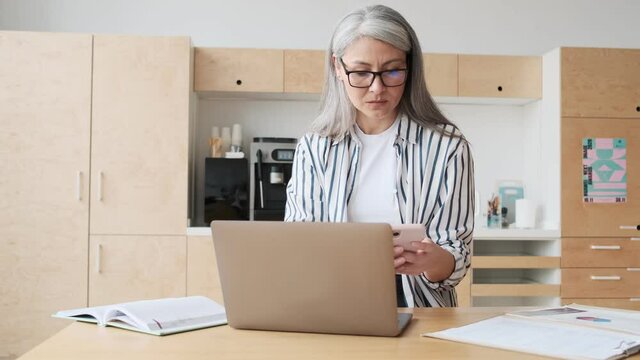 A serious elder business woman wearing eyeglasses is using her laptop computer while making notes in the kitchen at home