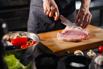 The male chef cuts raw meat with a knife on a wooden board. The restaurant kitchen cooking.