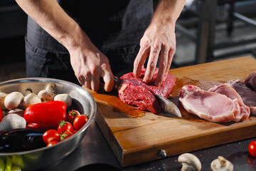The male chef cuts raw meat with a knife on a wooden board. The restaurant kitchen cooking.