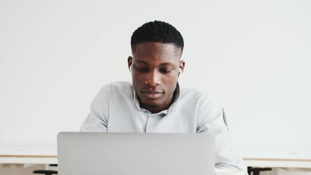 A happy black guy with earbuds is using his laptop sitting alone in the office