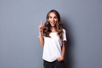 Delighted woman teenager with opened mouth looking in camera  holding up forefinger in white t-shirt on gray wall