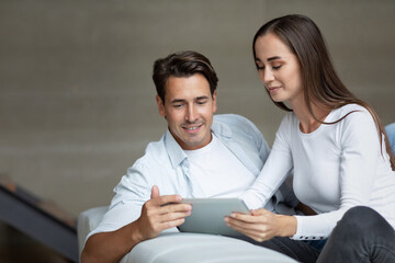 Smiling young couple sit relax on sofa in living room using modern gadgets together. Happy man and woman at home watch video on digital tablet.