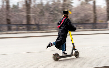 Plakat Happy Young woman in black graduation gown and black graduation cap with yellow tassel riding an electric scooter down street after graduation at university