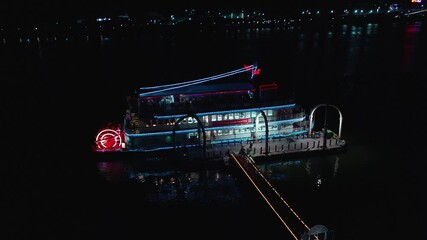 Aerial flight towards blue and red lighting restaurant boat ready for departure at night.