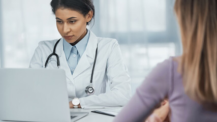 african american doctor in white coat using laptop near patient on blurred foreground