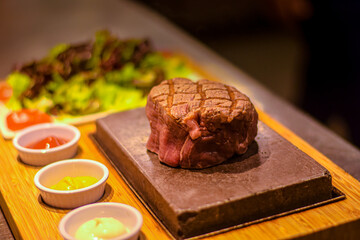 Grilled juicy beef steak smoking on hot stone served on wooden board with salad and sauces