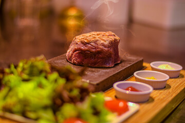 Grilled juicy beef steak smoking on hot stone served on wooden board with salad and sauces