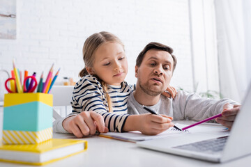 dad and daughter pointing at notebook near laptop on blurred foreground