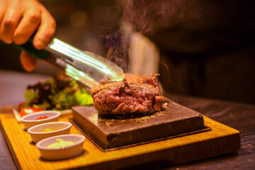 Chef turning grilled juicy beef steak smoking on hot stone served on wooden board with salad and sauces