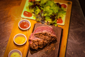 Grilled juicy beef steak smoking on hot stone served on wooden board with salad and sauces