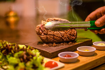 Chef turning grilled juicy beef steak smoking on hot stone served on wooden board with salad and sauces