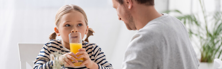 girl looking at father while holding glass of orange juice, banner