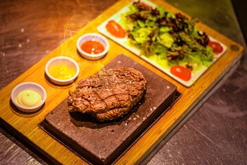 Grilled juicy beef steak smoking on hot stone served on wooden board with salad and sauces