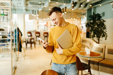 Young smiling business man freelancer in casual yellow sweater and jeans working remotely using laptop and phone sitting in cafe, selective focus