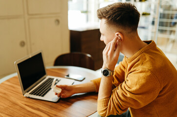 A young man is a freelance student studying learning working remotely online talking on the phone using wireless technologies headphones and modern gadgets in a public place in a cafe