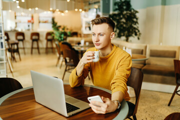 Young smiling business man freelancer in casual yellow sweater and jeans working remotely using laptop and phone sitting in cafe, selective focus