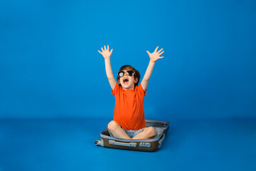 happy little boy wearing sunglasses sits in a suitcase on a blue background with space for text