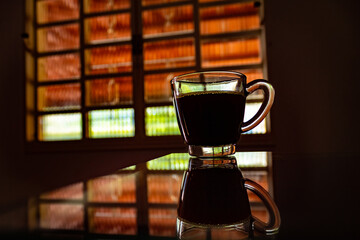 Uma xícara transparente com café sobre uma superfície reflexiva e uma janela ao fundo.