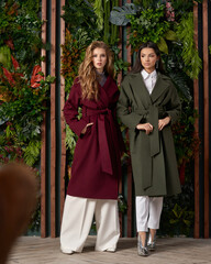 Two girls wearing colorful wool coats and posing against flowal wall. Blonde female model and brunette young woman with long hair standing. Fashion portrait