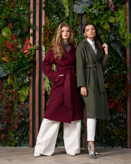 Two girls wearing colorful wool coats and posing against flowal wall. Blonde female model and brunette young woman with long hair standing. Fashion portrait