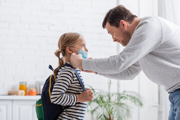 side view of man putting on medical mask on daughter with backpack