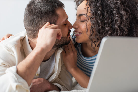 interracial couple kissing while chilling on bed near laptop