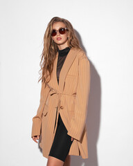 Young elegant teenager caucasian girl with long wavy blonde hair wearing black bike shorts, net blouse and oversize beige blazer standing and posing against white wall
